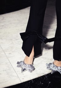 Pantalons noirs avec fentes sur les côtés, accentués par des nœuds en satin noir. Chaussures à talons hauts argentées et texturées avec de grands nœuds et des pointes.