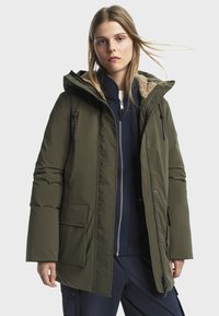 Zateplená parka v olivovo zelenej farbe so zapínaním na zips a gombíky, kožušinovou podšívkou kapucne, prednými vreckami a voľným strihom. Viditeľná textúrovaná vnútorná podšívka.