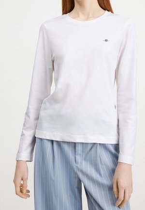 Vrouw draagt een effen wit shirt met lange mouwen met een klein logo op de borst, gecombineerd met lichtblauwe broek met fijne strepen.