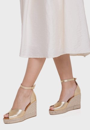 Jambes portant des sandales compensées métalliques dorées avec des brides à la cheville et des bouts ouverts, associées à une jupe blanche mi-longue.