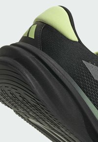 Zapatilla deportiva negra con detalles verdes, parte superior de malla texturizada y una suela de goma gruesa estriada para tracción y soporte.