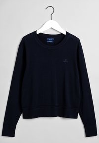 Maglione navy blue a girocollo con maniche lunghe, polsini e orlo a costine, appeso a un gancio bianco su uno sfondo grigio chiaro.