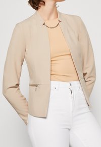 Beige tailored blazer with a smooth texture, structured design, and two front zippered pockets. Worn over a light top and white pants.