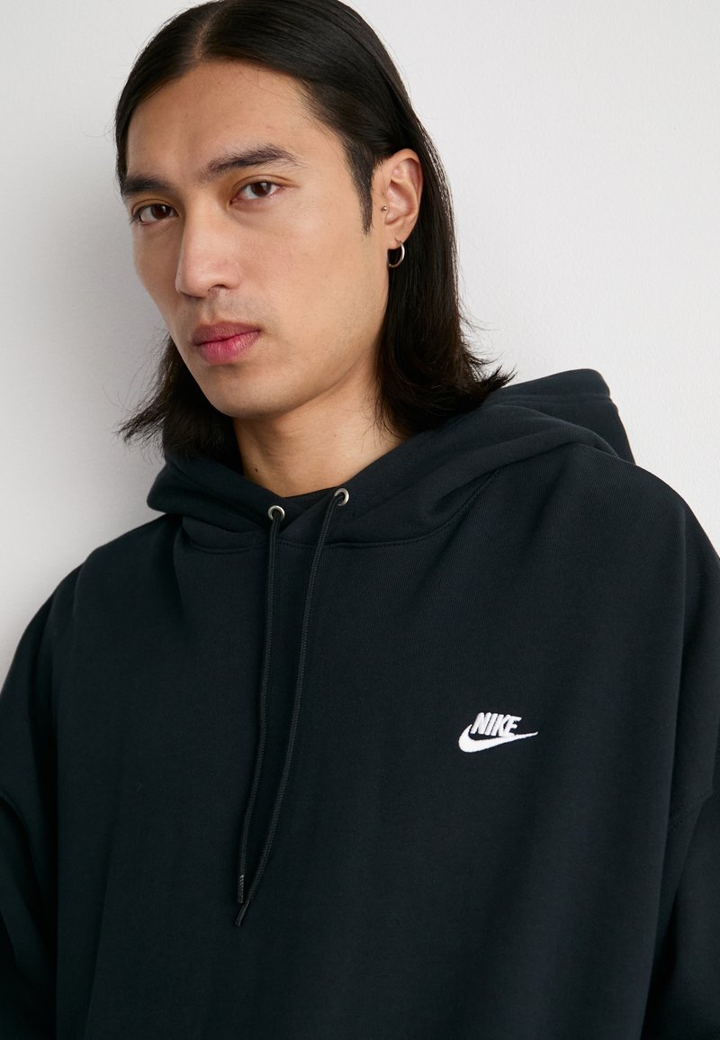 Homme Nike noir à capuche avec cordon de serrage, arborant un logo blanc sur la poitrine. Le tissu semble doux avec un design simple et décontracté.