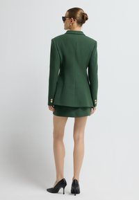 Vihreä räätälöity bleiseri, jossa on tiukka muotoilu, yhteensopiva minihame ja kultaiset koristepainikkeet. Mustat korkokenkäsaappaat, joissa on teksturoitu pinta.