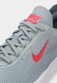Sive Nike Free tenisice s teksturiranim mrežnim gornjim dijelom, crvenim Nike logotipom i sivim vezicama. Sadrže podstavljen jezik i prozračan dizajn.