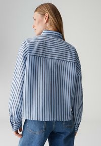 Chemise bleu clair à rayures avec une coupe décontractée, dotée d'un col, de poignets boutonnés, et d'un motif à rayures plus foncées contrastantes au dos.
