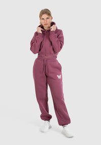 Felpa cropped con zip e pantaloni coordinati in morbido tessuto color bordeaux scuro. La felpa presenta un cappuccio con coulisse; i pantaloni hanno una stampa di farfalle bianche.