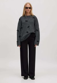 Grauer Pullover aus strukturiertem Stoff, mit schwarzen Schleifenakzenten, kombiniert mit einer schwarzen, weit geschnittenen Hose und schwarzen Schuhen. Minimalistisches Design.