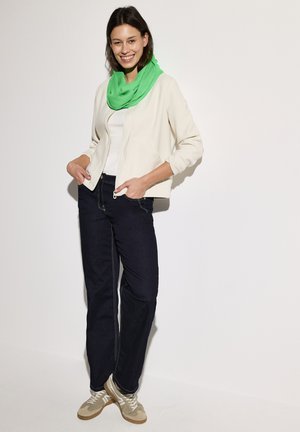 Licht beige zip-up jack, witte top, donkerblauwe jeans en groene sjaal. Model staat met handen in de zakken, terwijl ze tan en witte sneakers draagt.