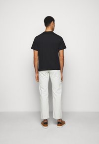 Camiseta de algodón negra de manga corta, combinada con pantalones de textura gris claro y zapatos marrones perforados, vista desde atrás.