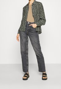Dunkelgrünes Hemd mit floralem Muster, beiges, figurbetontes Oberteil, hochgeschnittene, dunkle Jeans und schwarze Sandalen mit Riemen, die einen lockeren Schnitt und gerade Beine haben.