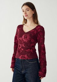 Burgundia pikad varrukad ülikond lillede tikandiga, V-kaelusega ja läbipaistvate varrukatega, koos tumedate teksadega.