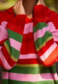 Pull rayé en rouge, rose, vert et orange. Avec une capuche et des poignets côtelés. Fabriqué en matière tricotée, mettant en avant de larges rayures horizontales.