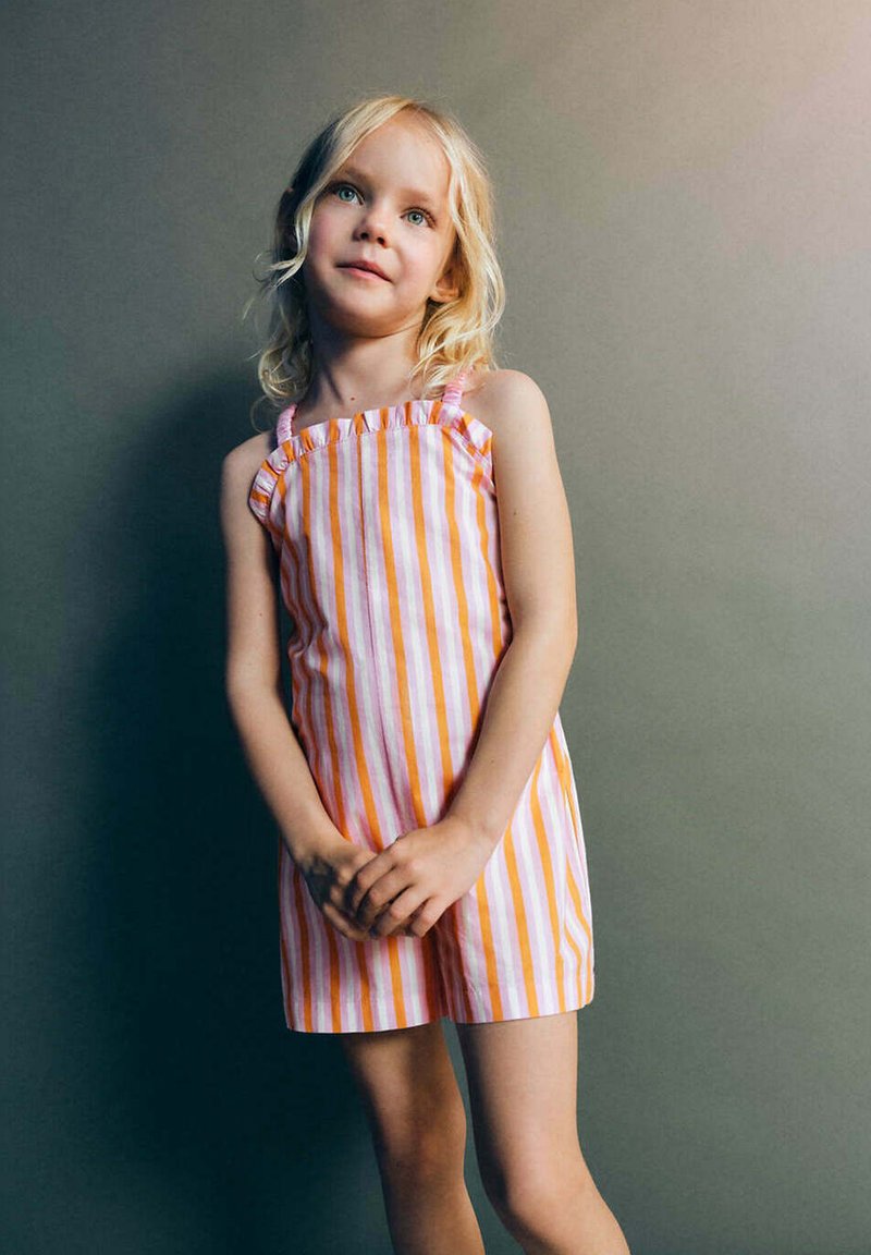 Jeune fille aux cheveux blonds portant une robe sans manches rayée rose, blanche et orange, debout devant un fond gris uni.