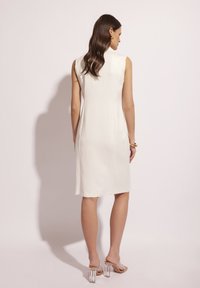 Biała sukienka bez rękawów o dopasowanym kroju, gładkiej fakturze i długości do kolan, zestawiona z przezroczystymi sandałami na obcasie. Prosty, elegancki design.