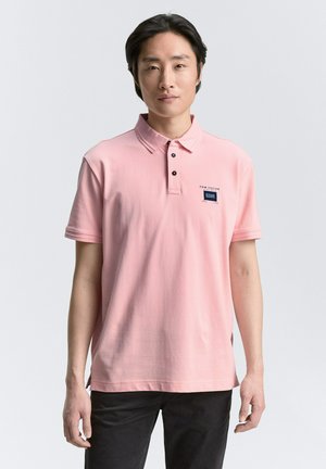 Polohemd aus leicht rosa Baumwolle mit einem Kragen, zwei schwarzen Knöpfen und einem kleinen marineblauen Logo auf der linken Brust. Kombiniert mit dunklen Hosen.
