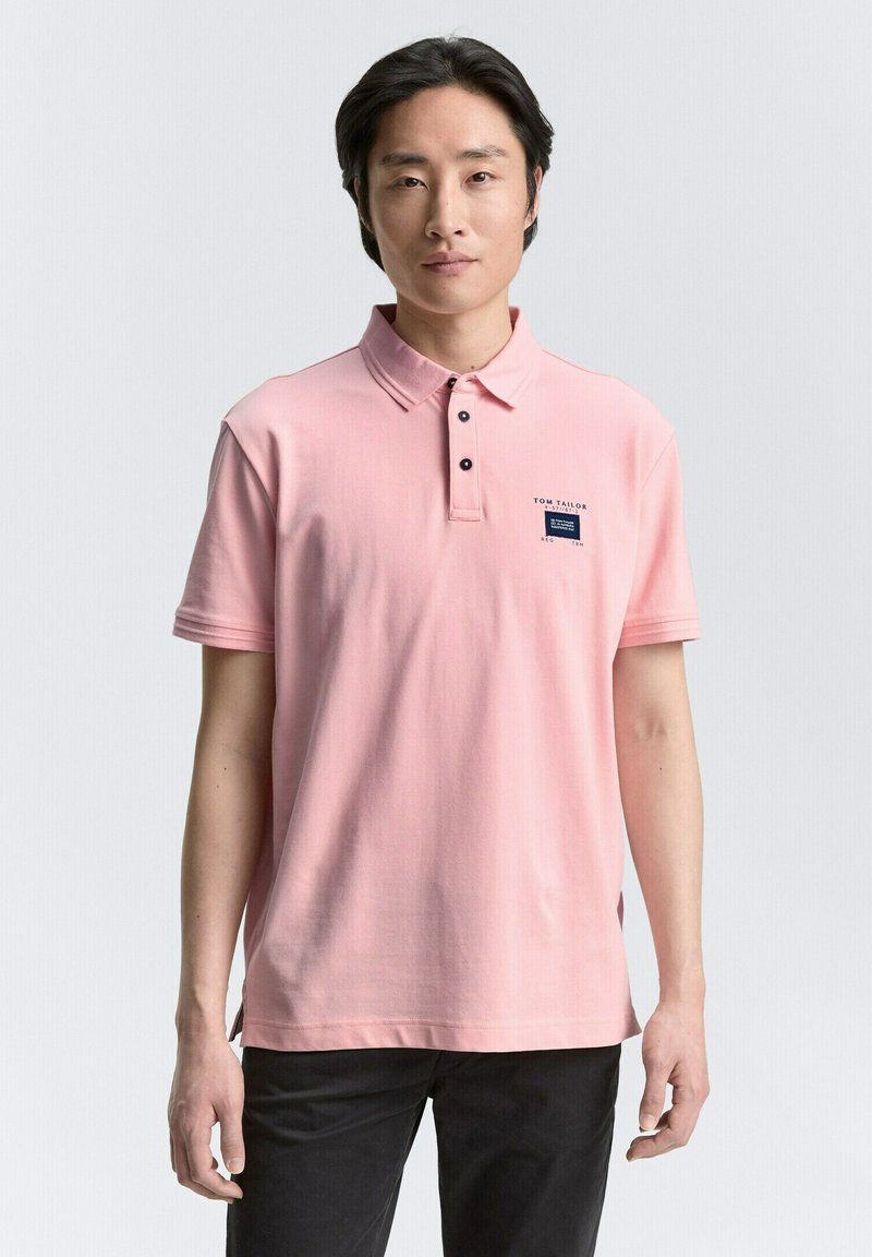 Camisa polo de algodón en color rosa claro con cuello, dos botones negros y un pequeño logo en azul marino en el lado izquierdo del pecho. Combinada con pantalones oscuros.