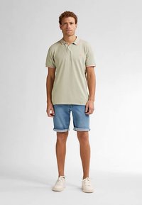 Vaaleanvihreä poolopaita, jossa on vaaleanpunaiset kauluskoristeet, yhdistetty farkkushortseihin. Mallilla on päällään kermaiset kengät ja hän seisoo yksinkertaisen taustan edessä.