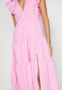 Różowa sukienka warstwowa z krótkimi falbankowymi rękawami, głębokim dekoltem w kształcie V i rozcięciem po boku. Wykonana z lekkiego materiału, z marszczonymi szwami dla uzyskania faktury.