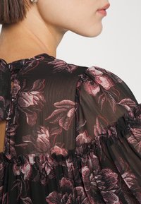 Blouse en tissu noir transparent avec un motif de roses et de feuilles dans des nuances de rouge. Caractéristiques des manches froncées et un détail d'ouverture en forme de clé au dos.