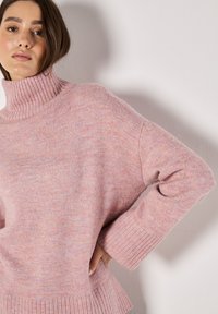 Hellrosafarbener Strickpullover mit hohem Rippkragen, weiten Ärmeln und einem strukturierten Muster, das Rippenakzente am Saum und an den Bündchen aufweist.
