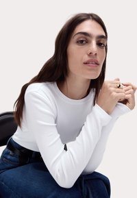 Langärmliges, weißes Rippenoberteil mit Rundhalsausschnitt, kombiniert mit blauer Jeans. Das Model hat lange braune Haare, trägt einen goldenen Ring und sitzt auf einem Stuhl.