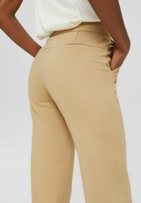 Pantalon beige à jambes larges avec une texture lisse et extensible. Dispose de poches latérales et d'une taille haute ajustée. Convient pour un usage décontracté ou professionnel.