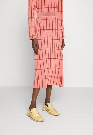 Mujer con una falda midi rosa y una blusa de manga larga a juego con un patrón de cuadrícula roja, combinados con mocasines beige sobre un suelo gris.