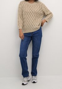 Pull en tricot beige avec un motif ondulé, associé à un jean bleu droit. Complété par des chaussures de sport grises et blanches.