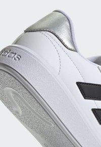 White sneaker with a textured surface, black accents, and a shiny silver heel. Features a rubber outsole and stitched detailing.