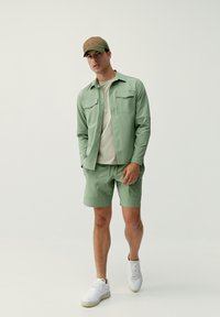 Ljust grön skjorta med knappärmar och två bröstfickor, beige t-shirt, matchande gröna shorts och en brun och grön keps. Vita sneakers.