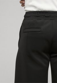 Personne portant un pantalon noir à taille élastique avec une poche passepoilée au dos, associé à un haut blanc rentré, debout devant un fond uni.