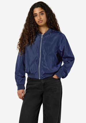 Giovane donna con lunghi capelli ricci che indossa una giacca bomber blu navy con la cerniera e jeans neri, in piedi con una mano in tasca.