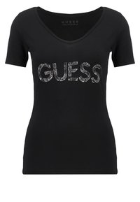 Camiseta negra de manga corta con escote en V, que presenta un logo brillante "GUESS" en lentejuelas en el pecho. Tela suave y lisa.