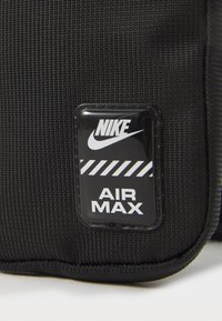 Geantă din nylon texturat negru, cu un patch lucios cu logo-ul Nike negru și dungi albe și textul "AIR MAX" dedesubt. Formă rectangulară.