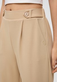 Beige hoog-getailleerde broeken met plooien, voorzien van een knoopsluiting en een klein accentlabel op de voorkzak. Zachte, gestructureerde stof.