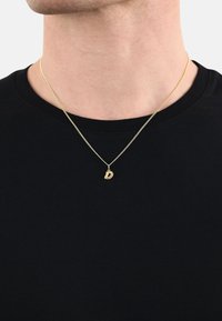 Collana in tono oro con un piccolo pendente a forma di lettera "D" appeso a una sottile catena, indossata sopra una t-shirt nera a girocollo.