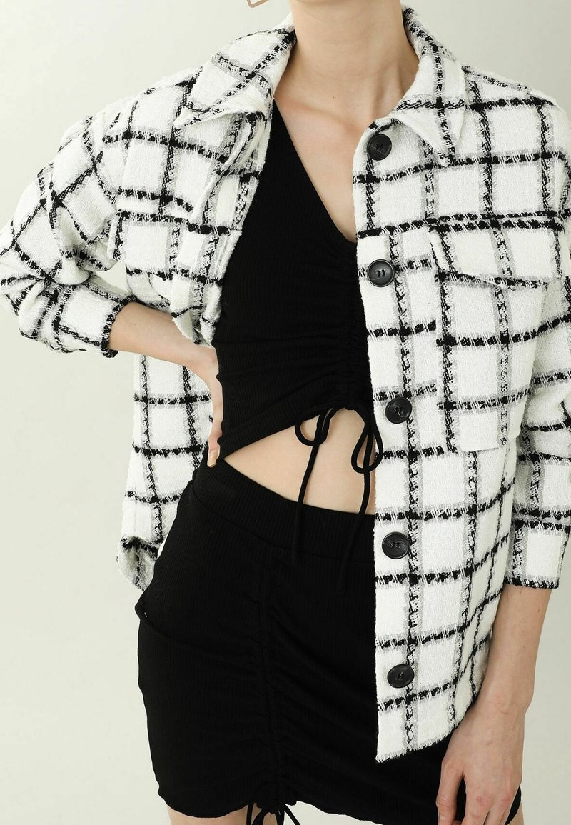 Biała i czarna kurtka w kratę wykonana z teksturowanej tkaniny, z długimi rękawami, zapięciem na guziki oraz dwoma przednimi kieszeniami, noszona na czarnym stroju.