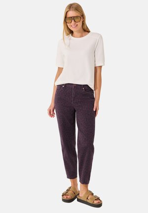Donna che indossa una maglietta bianca a maniche corte, pantaloni viola scuro con stampa leopardata, sandali platform color cuoio e grandi occhiali da sole con lenti gialle, sorridente con le mani rilassate.