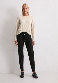 Eine Frau steht vor einer weißen Wand und trägt einen cremefarbenen Strickpullover, schwarze Jogginghosen mit Kordelzug und schwarze Slipper. Sie schaut nach vorne, und ihre Hände sind entspannt.