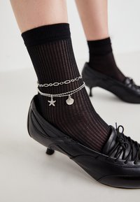 Calzino nero a coste indossato con una scarpa di pelle nera con tacco basso. È visibile un braccialetto alla caviglia in argento con un charm a forma di stella marina e conchiglia.