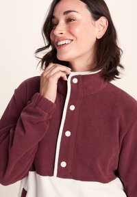 Smiling woman zipping up a maroon and white fleece jacket with snap buttons on a light background.