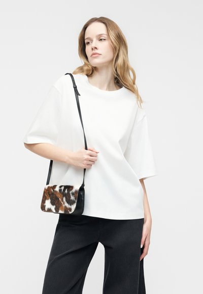 Γυναίκα που φοράει χαλαρό λευκό μπλουζάκι και μαύρο παντελόνι, κρατώντας μια μικρή τσάντα ώμου με καφέ και λευκή φλοράλ επιφάνεια με ζωικό μοτίβο.