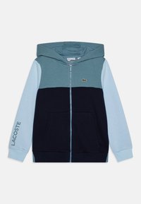 Sweatshirt zippé à capuche bleu et marine, présentant un design en color-block, des poches avant et un logo Lacoste vert sur la poitrine gauche.