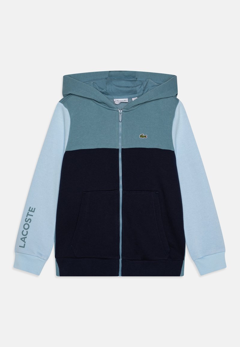 Sweatshirt zippé à capuche bleu et marine, présentant un design en color-block, des poches avant et un logo Lacoste vert sur la poitrine gauche.