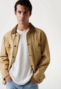 Jonge man met donker haar in een beige jas over een wit shirt en lichtblauwe spijkerbroek, met handen in zijn zakken, staande tegen een witte achtergrond.