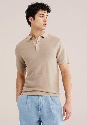 Młody mężczyzna z kręconymi włosami, ubrany w beżową koszulkę polo z krótkim rękawem i jasnoniebieskie dżinsy, patrzy w lewo z rękami w kieszeniach.