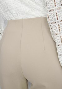 Pantalones beige de cintura alta en un tejido suave y texturizado con un detalle de costura discreto, combinados con una blusa blanca de encaje con un patrón intrincado.