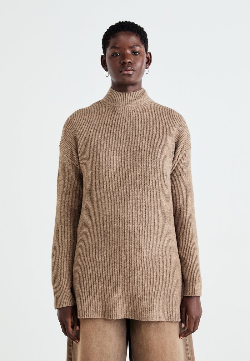 Pull en maille côtelée beige à col roulé avec des manches longues et des fentes sur les côtés, présentant une coupe décontractée et une texture douce.